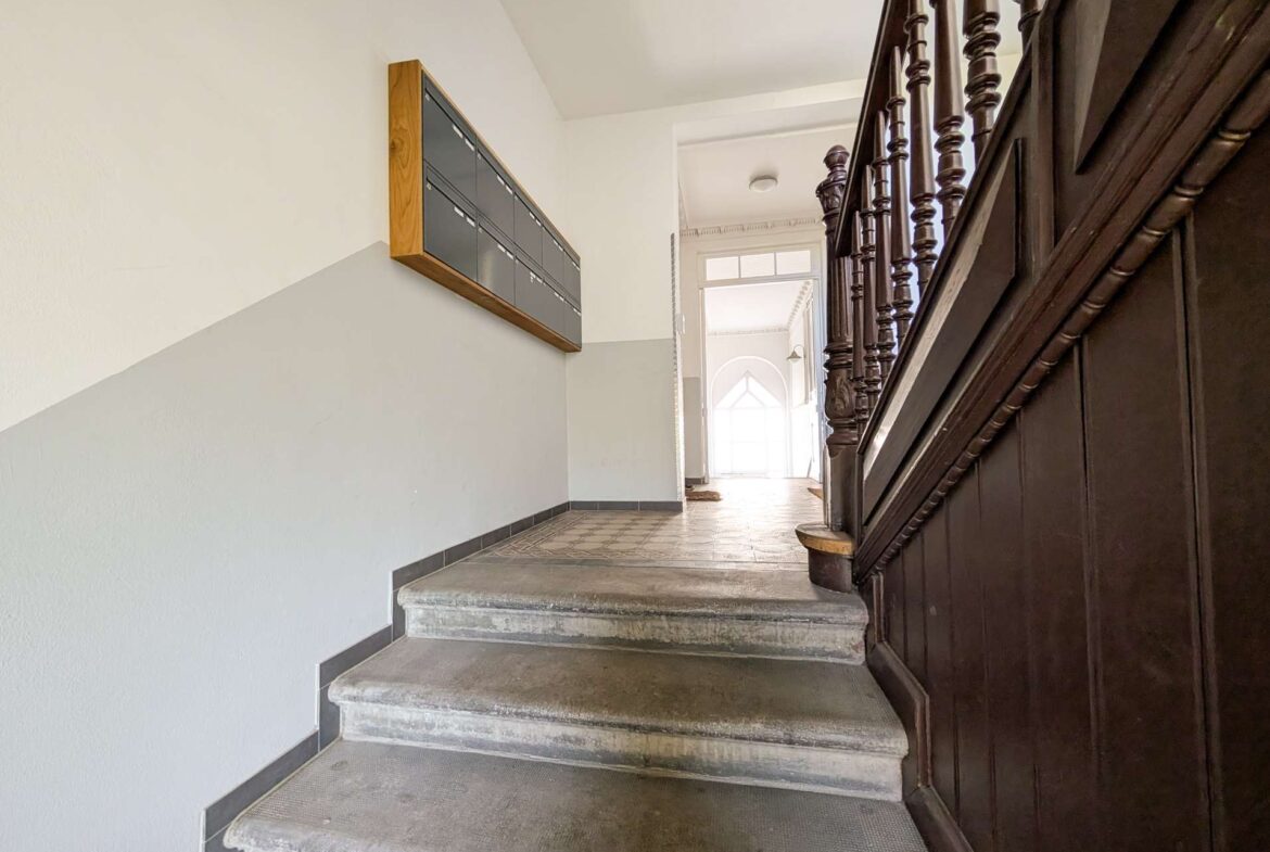 Interior stairwell with grey walls, row of black mailboxes on the left, and dark wooden banisters on the right leading to a bright doorway.
