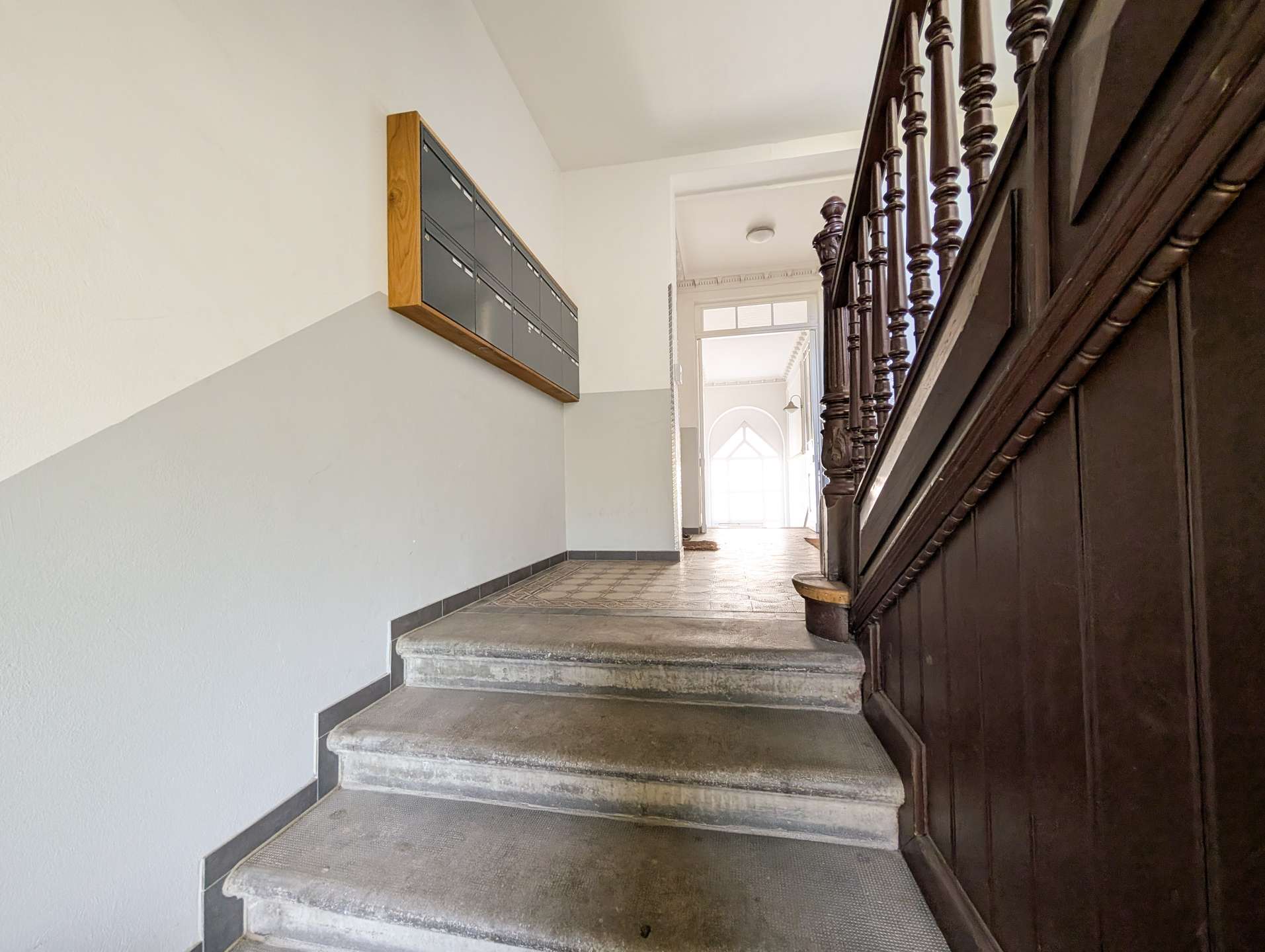 Interior stairwell with grey walls, row of black mailboxes on the left, and dark wooden banisters on the right leading to a bright doorway.