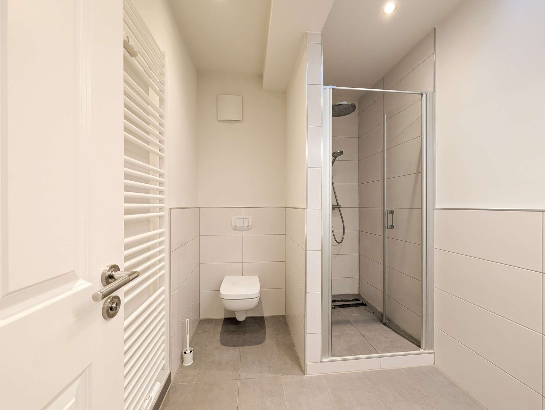 Small modern bathroom with wall-hung toilet, white towel warmer on the left, and a glass-door shower enclosure on the right.