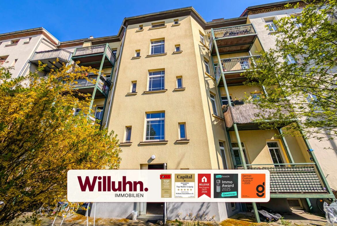 Four-story apartment building with beige facade and green balconies under a blue sky; prominent Willuhn Immobilien sign at street level.