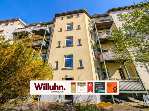 Four-story apartment building with beige facade and green balconies under a blue sky; prominent Willuhn Immobilien sign at street level.