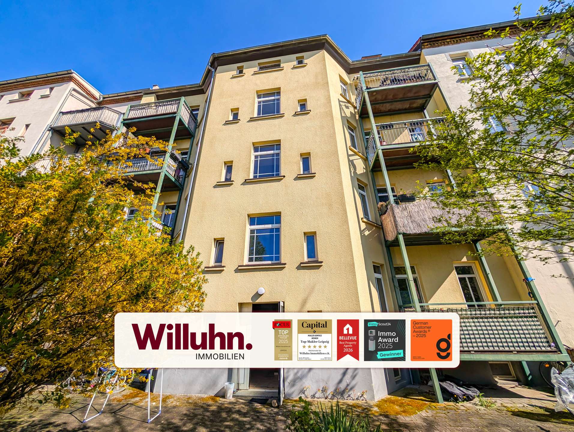 Four-story apartment building with beige facade and green balconies under a blue sky; prominent Willuhn Immobilien sign at street level.