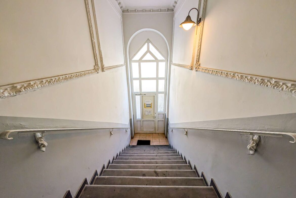 Narrow stairwell with decorative molding on pale walls, black metal handrails, and a large arched window at the landing ahead.