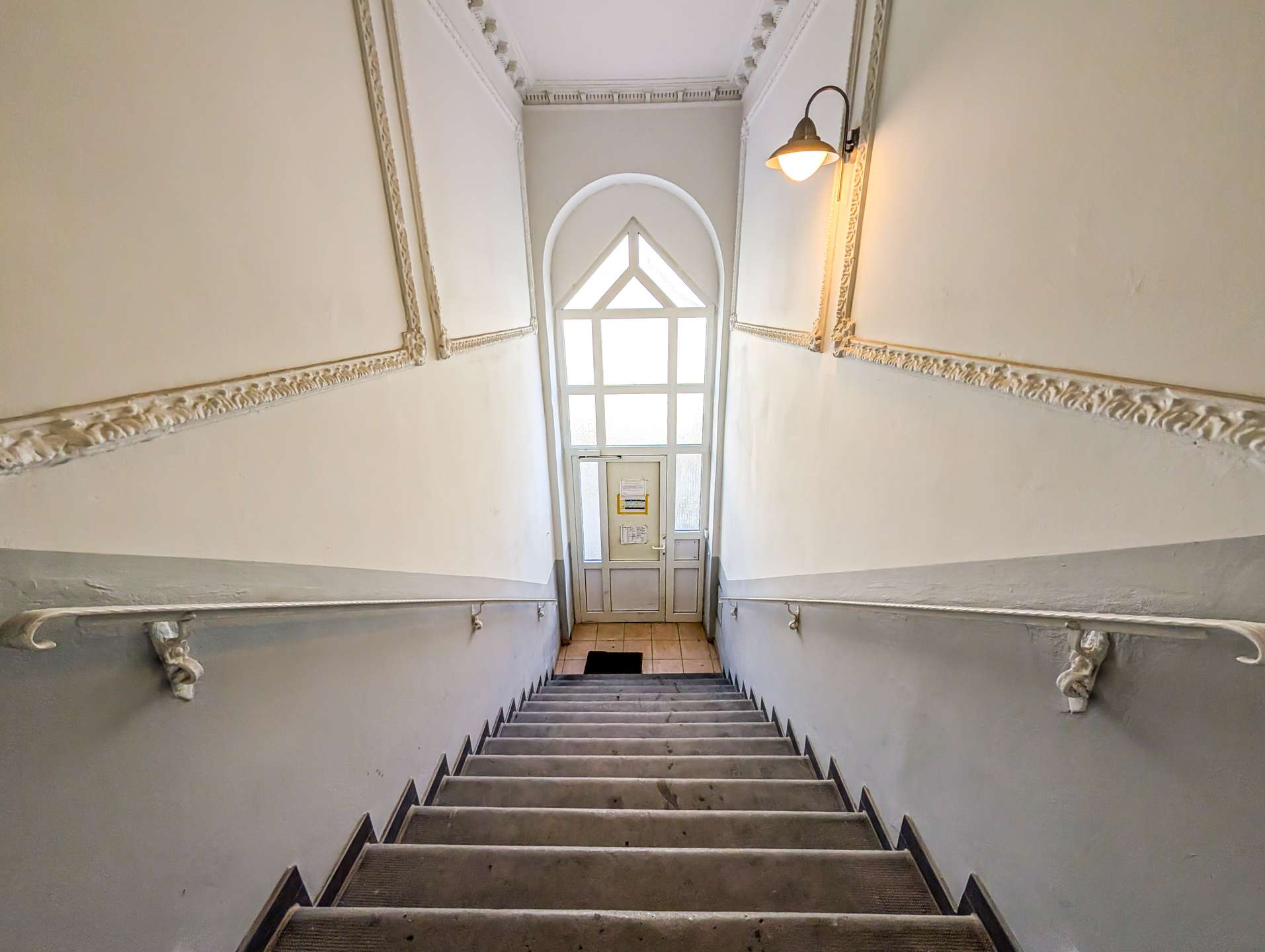 Narrow stairwell with decorative molding on pale walls, black metal handrails, and a large arched window at the landing ahead.
