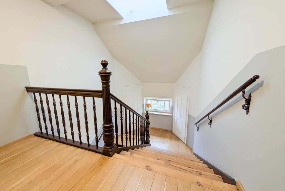 Interior stairwell with a dark wooden railing, turned balusters, and light wood stairs beneath a bright skylight in a white-walled space.