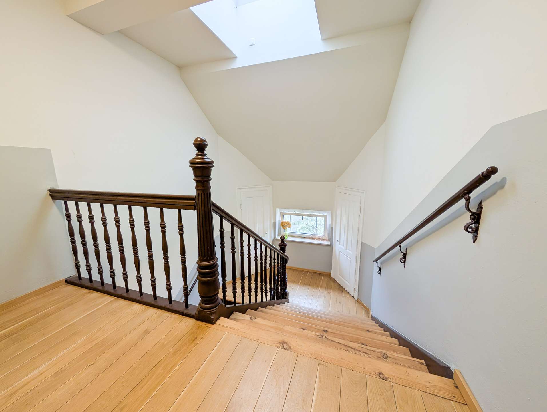 Interior stairwell with a dark wooden railing, turned balusters, and light wood stairs beneath a bright skylight in a white-walled space.