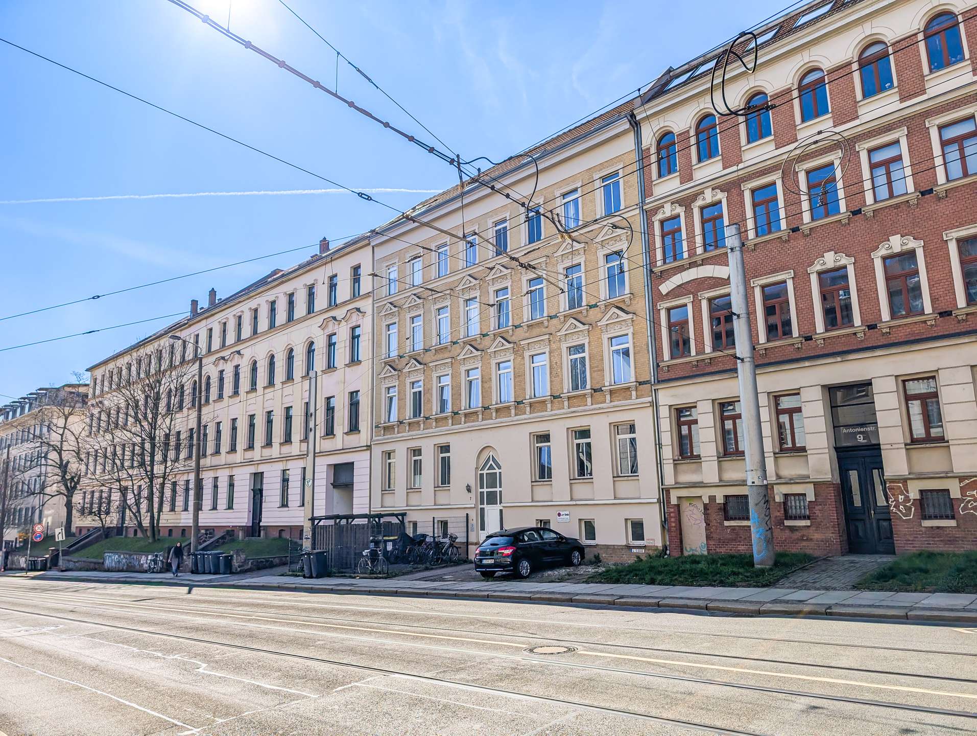 Row of beige and red brick historic apartment buildings along a tram line with overhead wires on a sunny day.