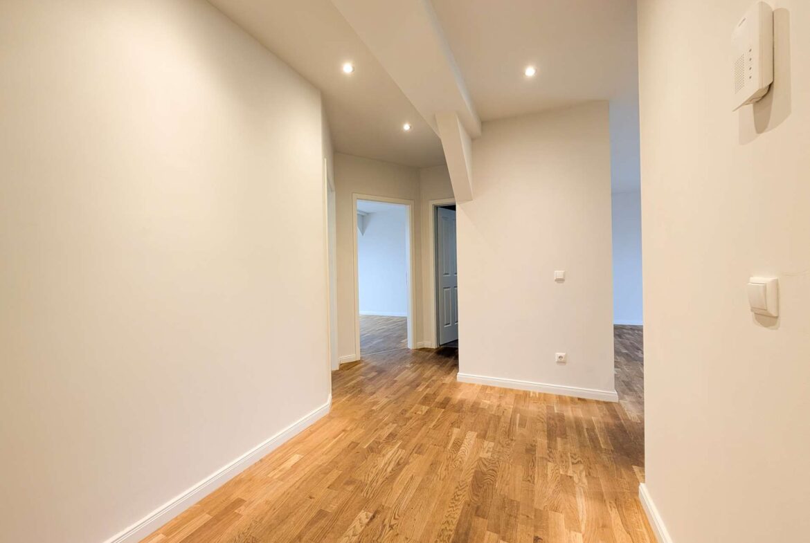 Narrow hallway with light wood flooring, white walls, and recessed ceiling lights; doorways at the end.