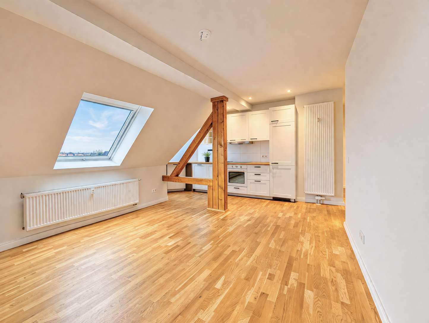 Bright attic with sloped white ceilings, skylight window, and a wooden beam support; open-plan kitchen with white cabinets and stainless appliances; light hardwood floor, radiator along the wall.