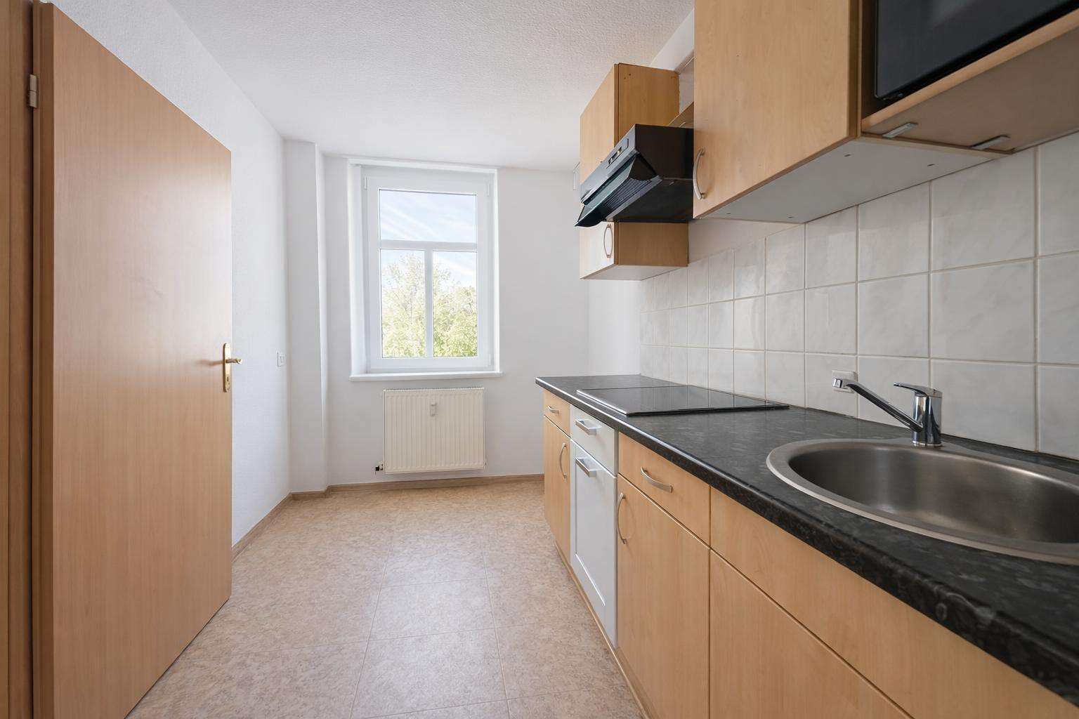 Compact kitchen with light wood cabinets, black countertop, stainless sink, electric stovetop, tiled backsplash, a window and radiator visible in a bright, narrow space.