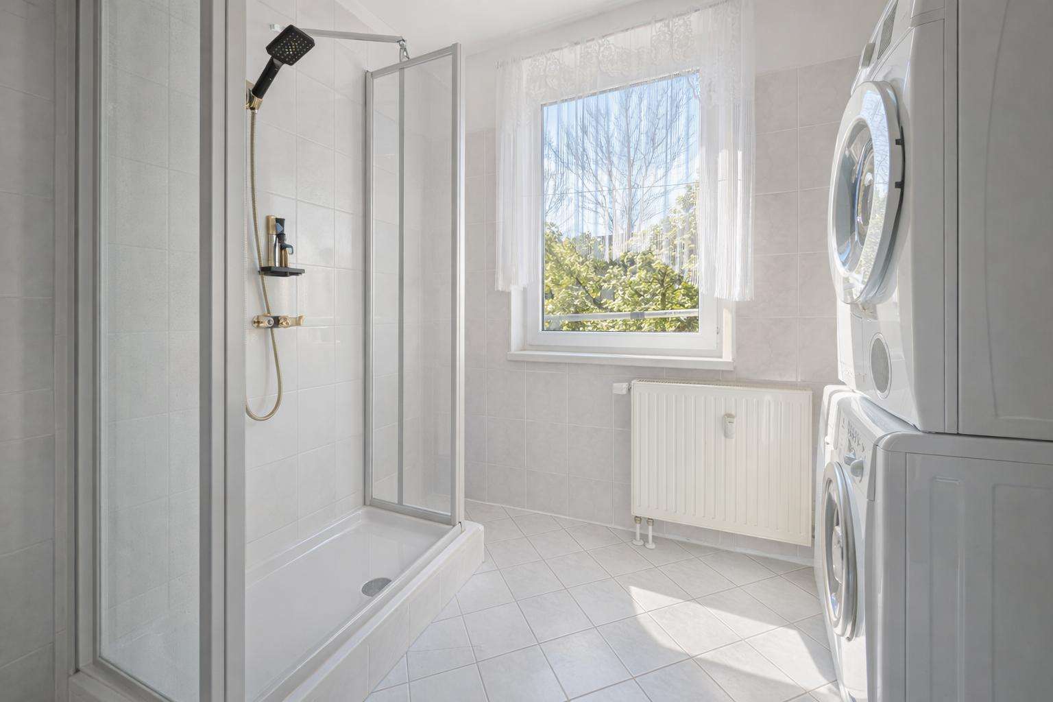Bright bathroom with a glass-enclosed shower, stacked washer and dryer on the right, and a window with lace curtains.