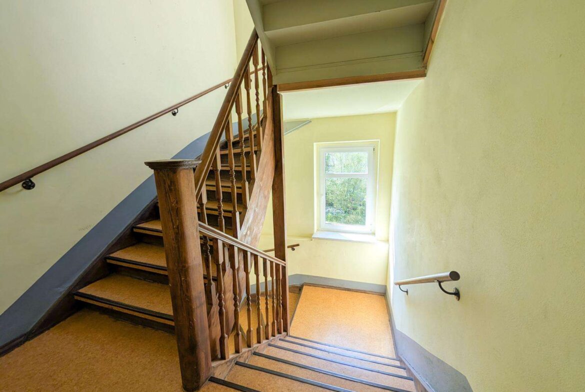 Wooden staircase in a pale yellow stairwell with a window letting in daylight. A dark wood banister runs along the stairs.