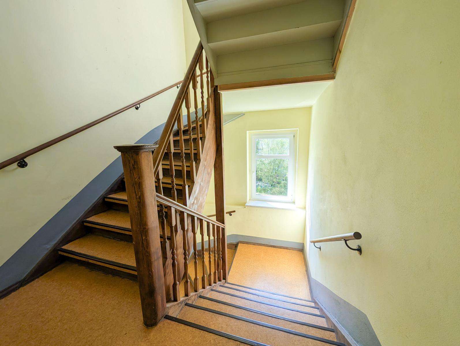 Wooden staircase in a pale yellow stairwell with a window letting in daylight. A dark wood banister runs along the stairs.