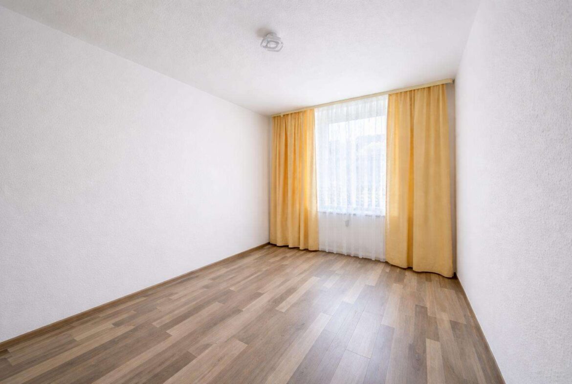 Empty rectangular room with light wood laminate flooring and a large window dressed in sheer white curtains plus mustard-yellow drapes.