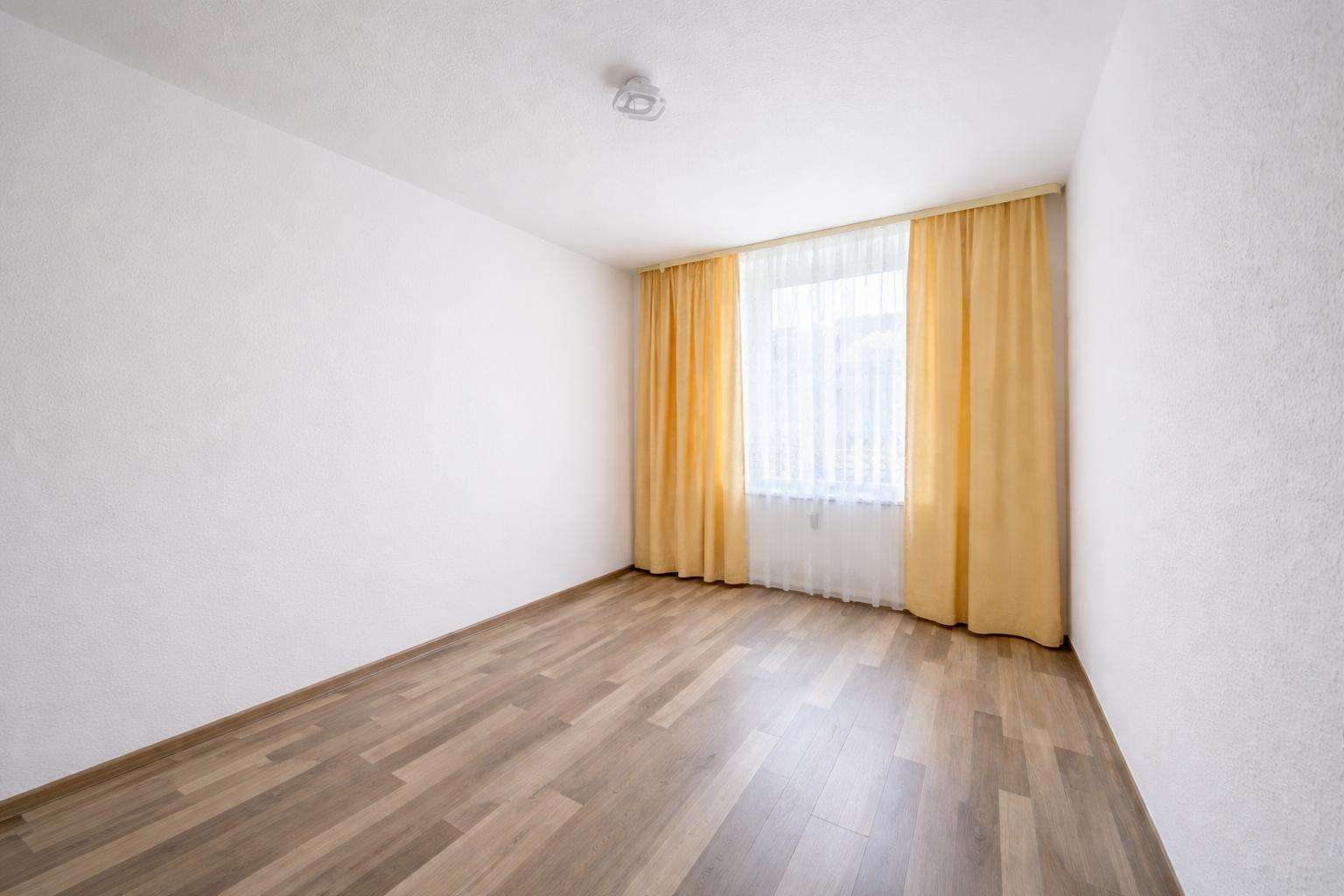 Empty rectangular room with light wood laminate flooring and a large window dressed in sheer white curtains plus mustard-yellow drapes.