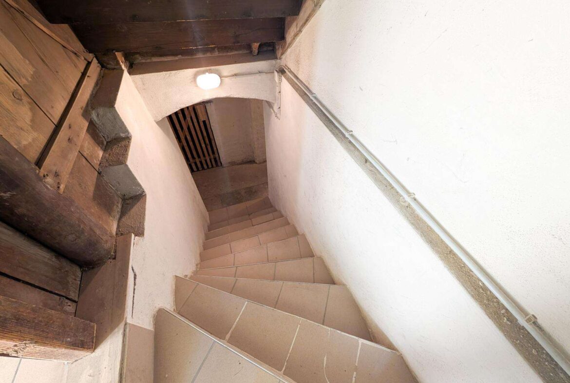 Basement stairwell with beige tiled steps, white walls, a metal handrail on the right, and a round ceiling light.