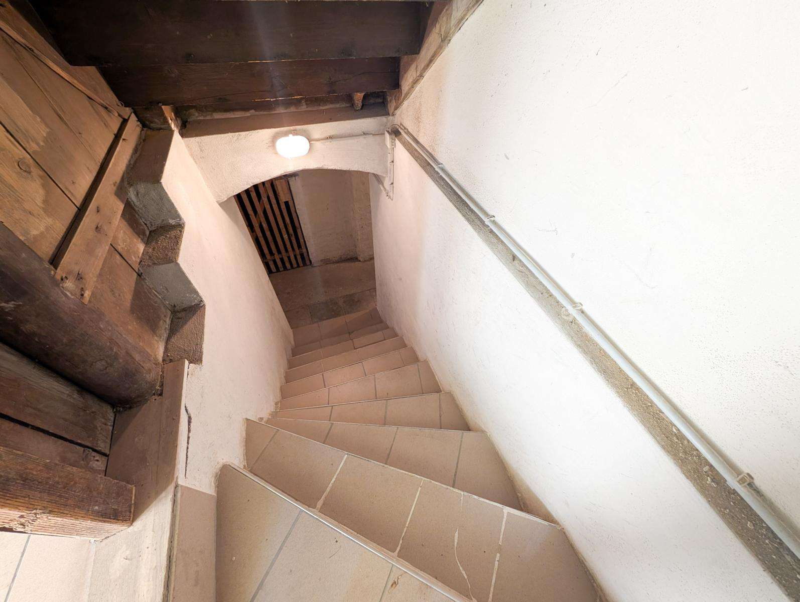 Basement stairwell with beige tiled steps, white walls, a metal handrail on the right, and a round ceiling light.