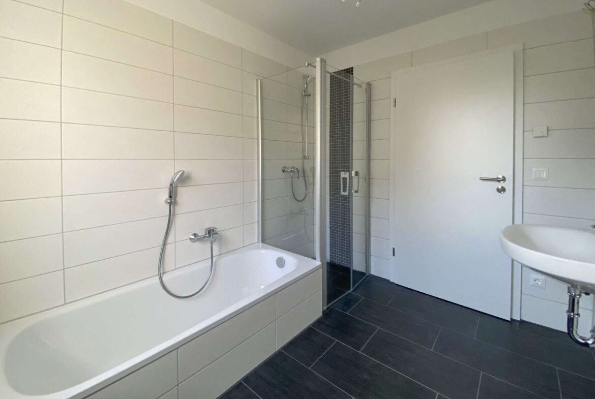 White bathtub along the left with a handheld shower, adjacent glass shower enclosure, and white tiled walls; dark floor tiles extend across the room.