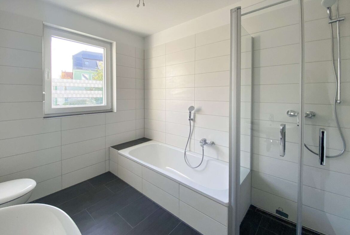 Modern bathroom with white tiled walls, built-in bathtub, glass-enclosed shower, window and dark floor tiles (toilet partially visible at left).