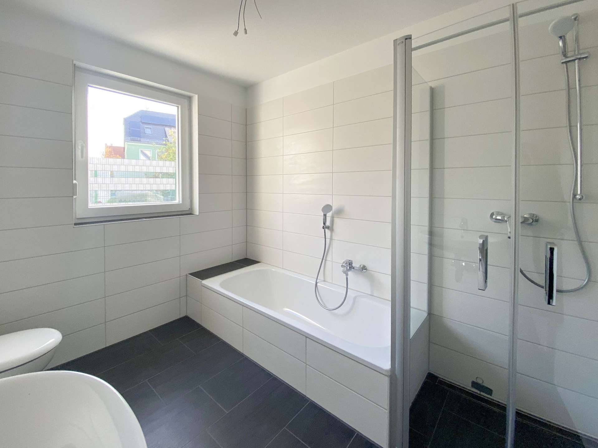 Modern bathroom with white tiled walls, built-in bathtub, glass-enclosed shower, window and dark floor tiles (toilet partially visible at left).