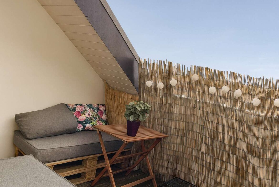 Cozy outdoor balcony corner with a gray cushioned sofa, floral pillow, wooden folding table, and a potted plant, next to a reed privacy screen with string lights against a blue sky.