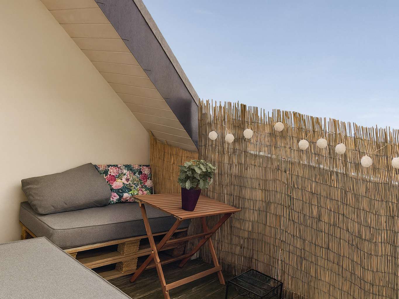 Cozy outdoor balcony corner with a gray cushioned sofa, floral pillow, wooden folding table, and a potted plant, next to a reed privacy screen with string lights against a blue sky.