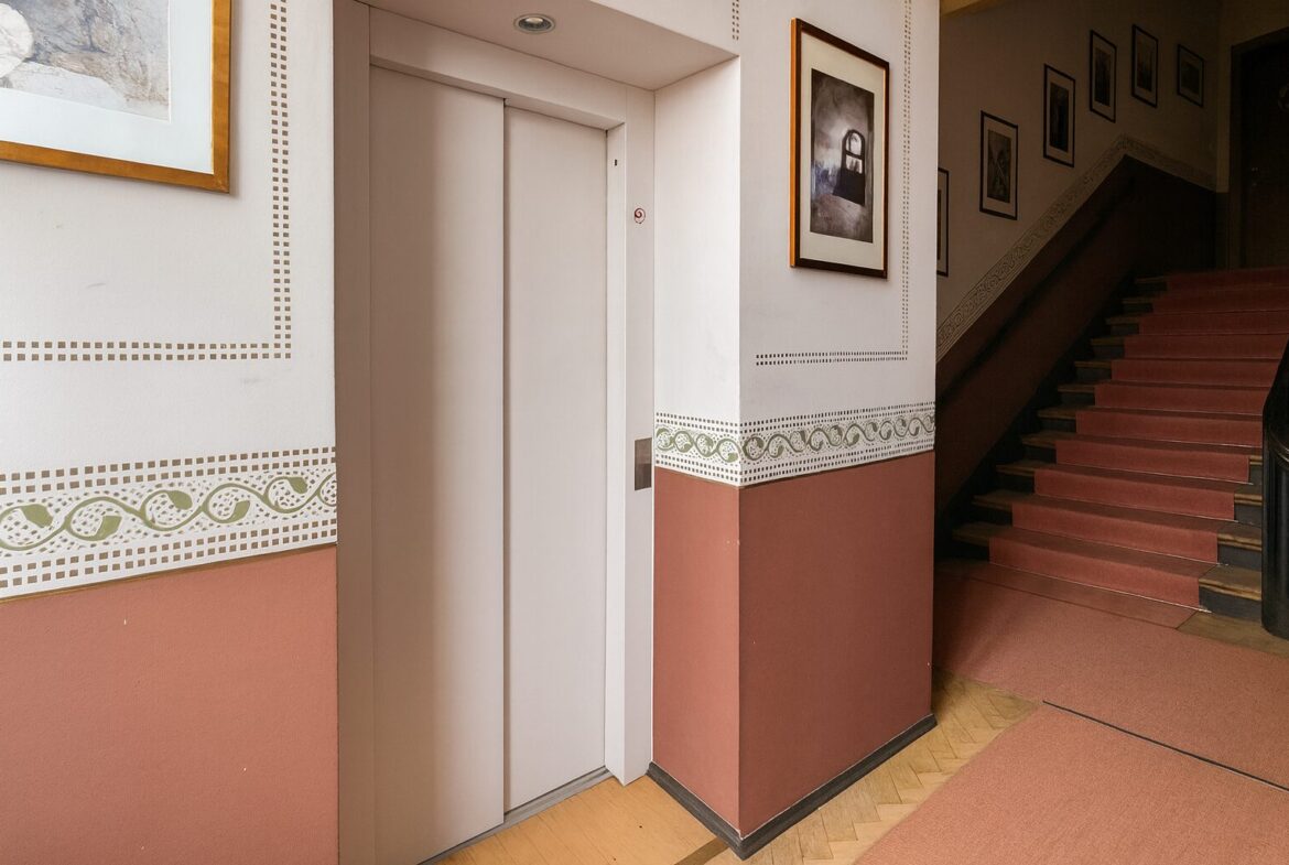 Elevator doors in a decorative hallway with framed art and a staircase to the right.