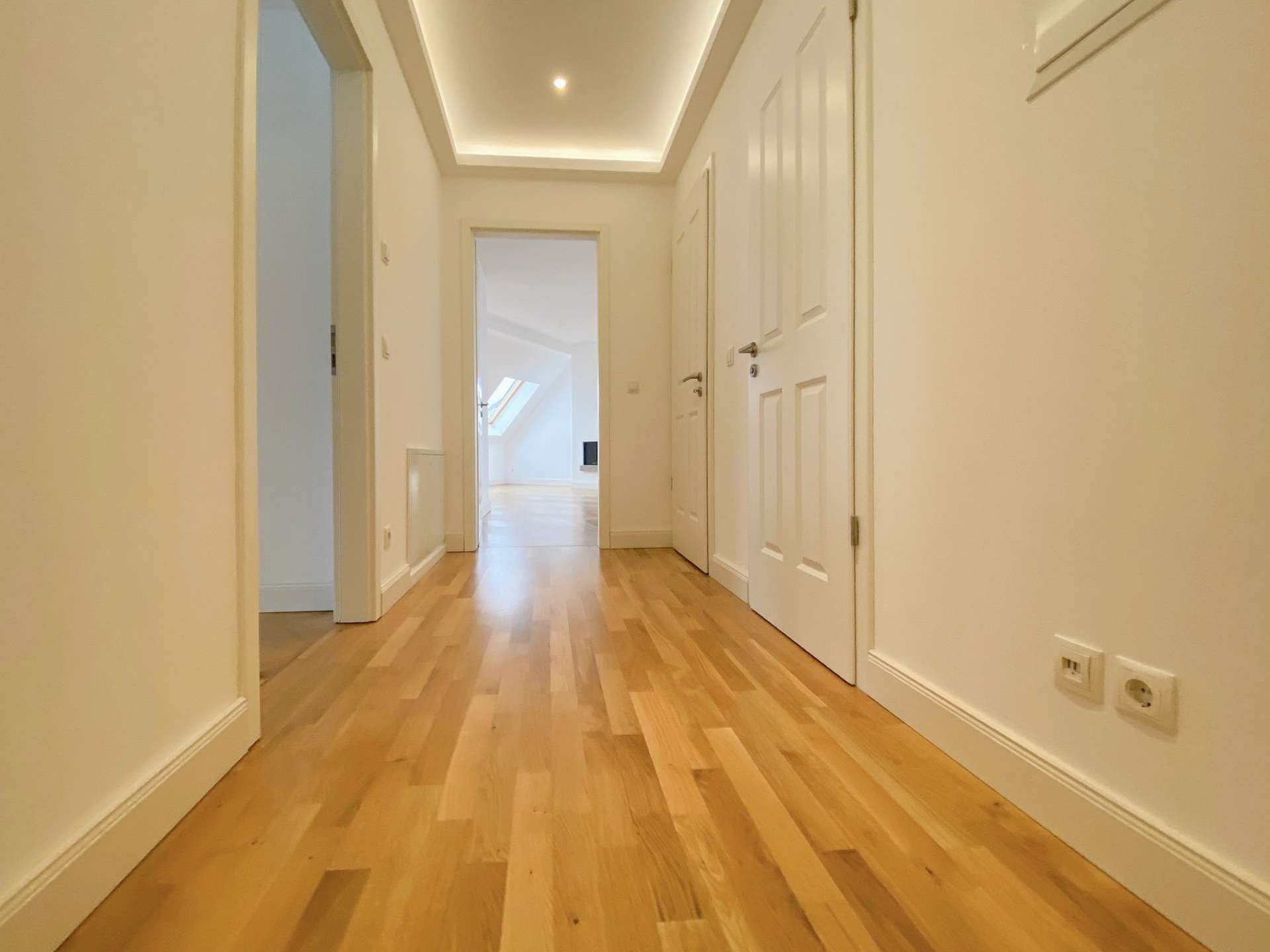 Bright white hallway with light wood flooring and white doors, leading to a sunlit room.