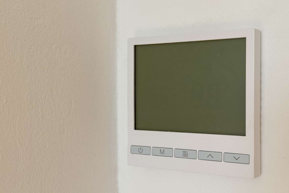 White wall-mounted thermostat with a blank greenish LCD screen and five light-gray control buttons below
