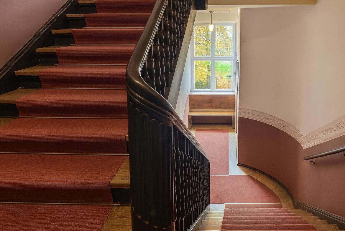Interior staircase with red carpet and dark wooden banister, leading to a landing with a large window showing trees outside.