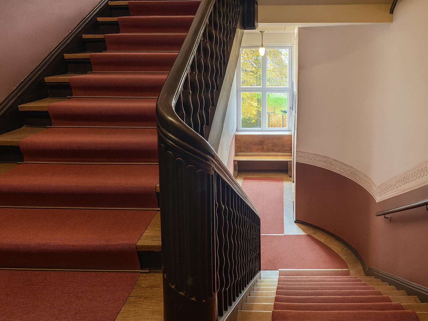 Interior staircase with red carpet and dark wooden banister, leading to a landing with a large window showing trees outside.