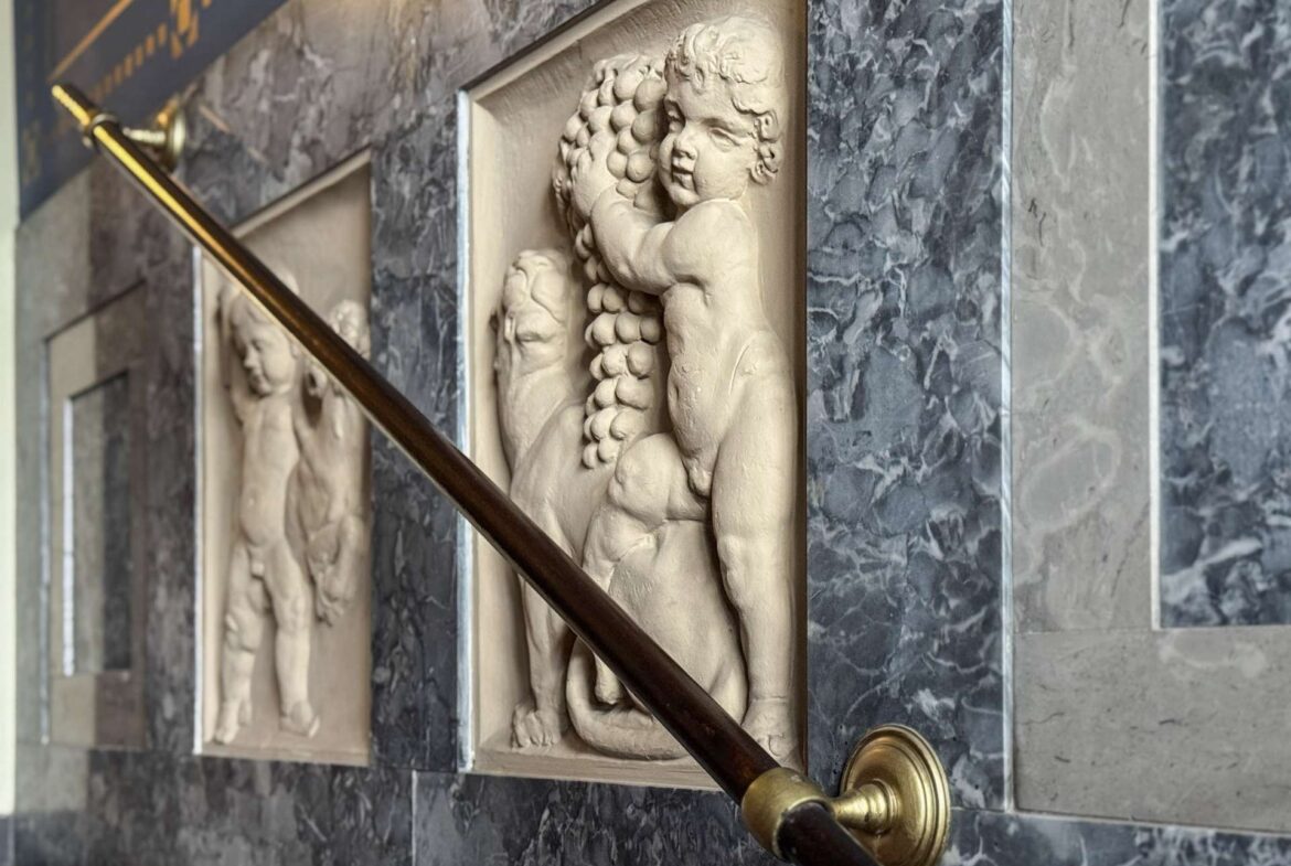 Marble wall with rectangular relief panels featuring child figures and a brass handrail in the foreground.