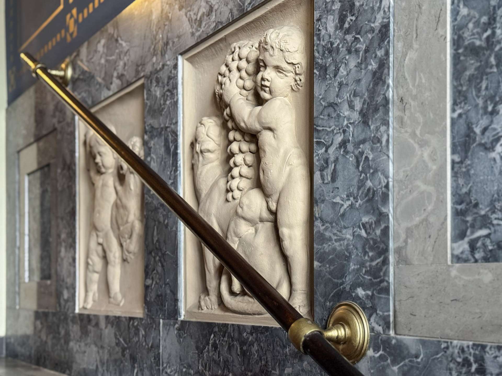 Marble wall with rectangular relief panels featuring child figures and a brass handrail in the foreground.