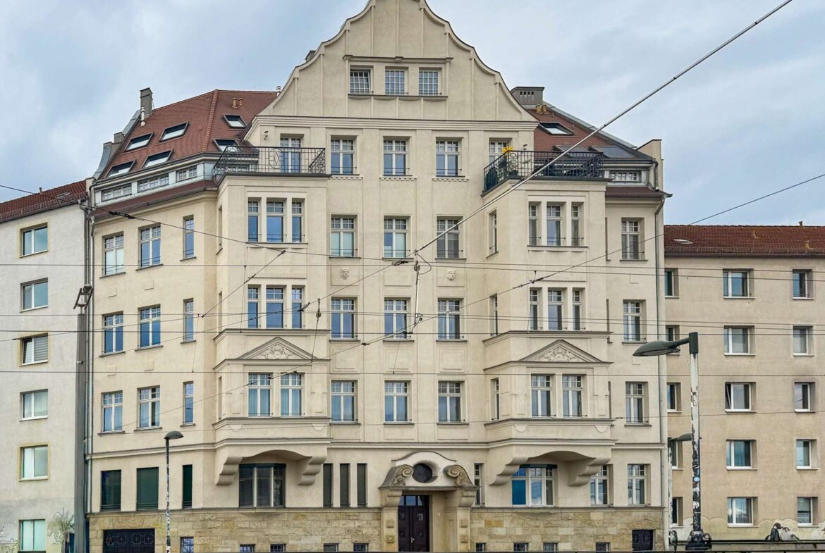 Elegant beige apartment building with ornate facade, arched entrance, balconies, and red-tiled roof seen behind tram wires.