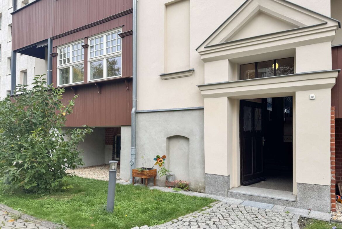 Exterior view of a residential building entrance: cream facade, open black door, with a small garden and wooden planter beside a windmill decoration.