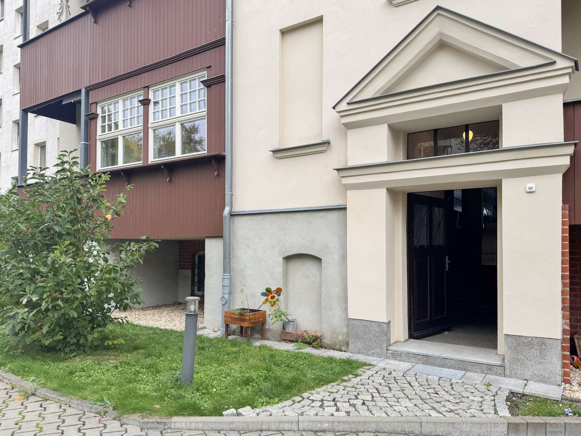 Exterior view of a residential building entrance: cream facade, open black door, with a small garden and wooden planter beside a windmill decoration.