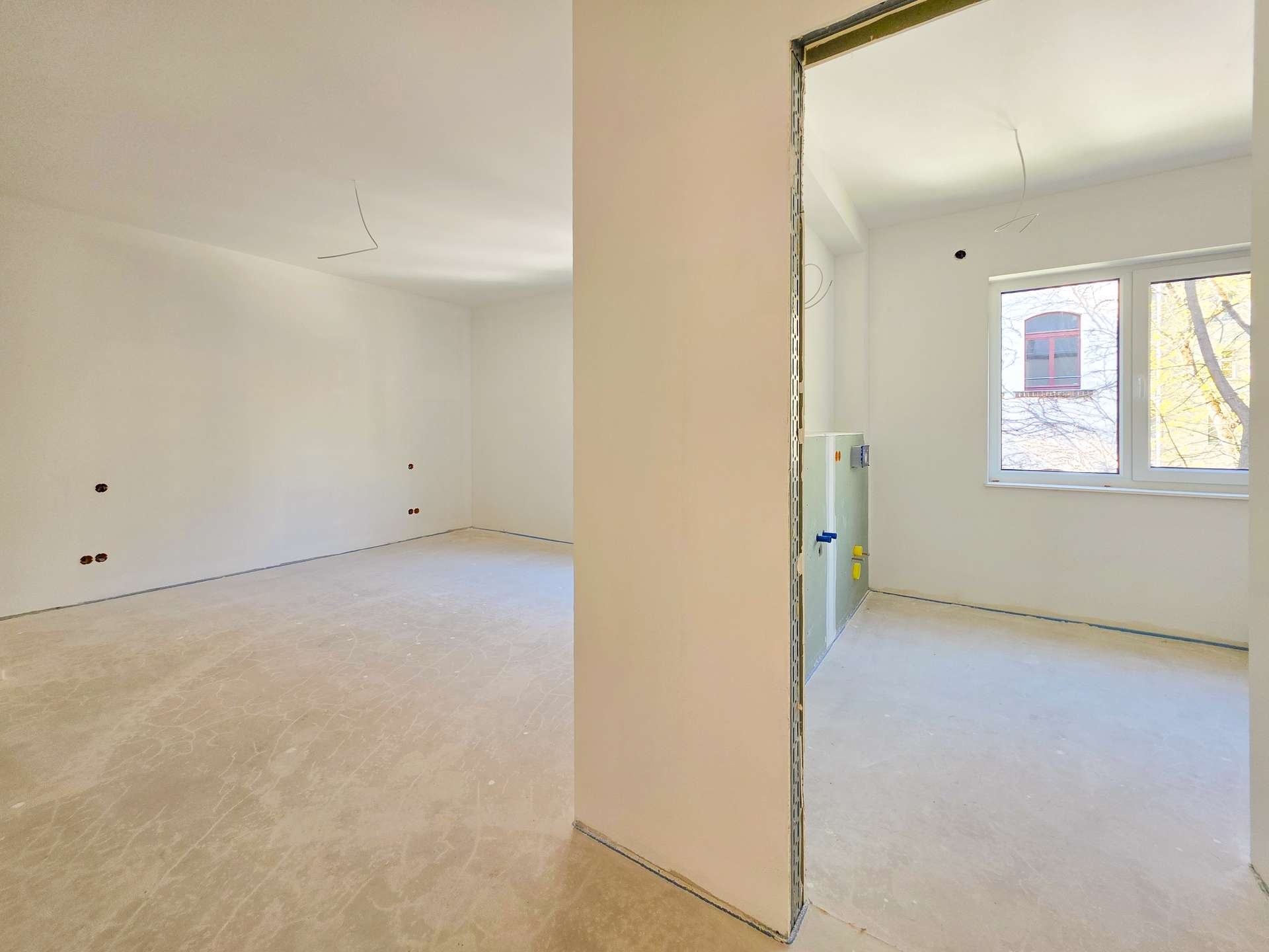 Open-plan unfinished room with exposed electrical wiring from ceiling and wall, white walls, and a large window letting in daylight.