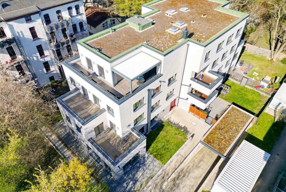 Aerial view of a white modern apartment complex with green-edged flat roofs, balconies, and a grassy courtyard area surrounded by trees.