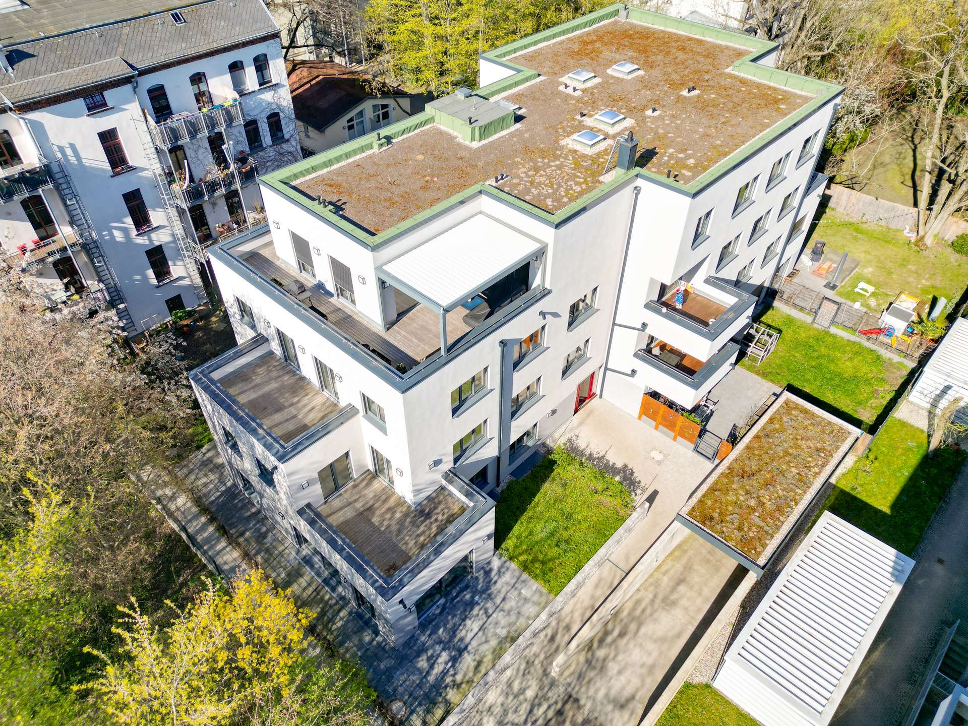 Aerial view of a white modern apartment complex with green-edged flat roofs, balconies, and a grassy courtyard area surrounded by trees.