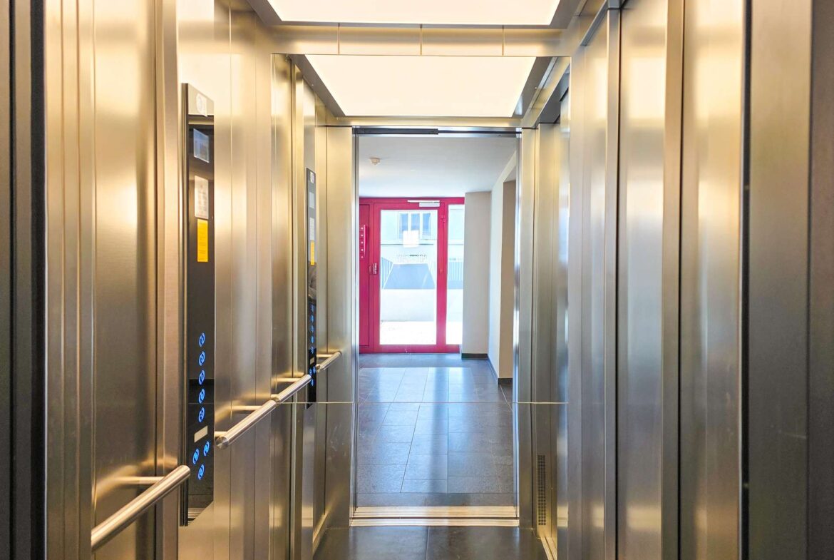 Elevator cabin with stainless steel walls and blue-lit control panel, doors open to a bright hallway ending at a red-framed door.