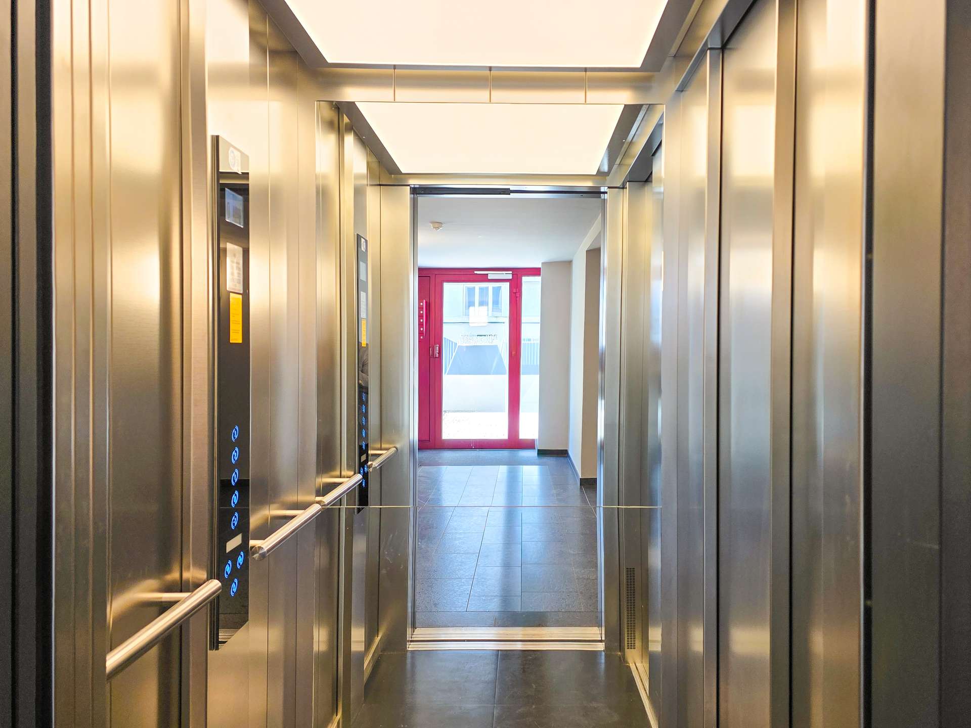 Elevator cabin with stainless steel walls and blue-lit control panel, doors open to a bright hallway ending at a red-framed door.
