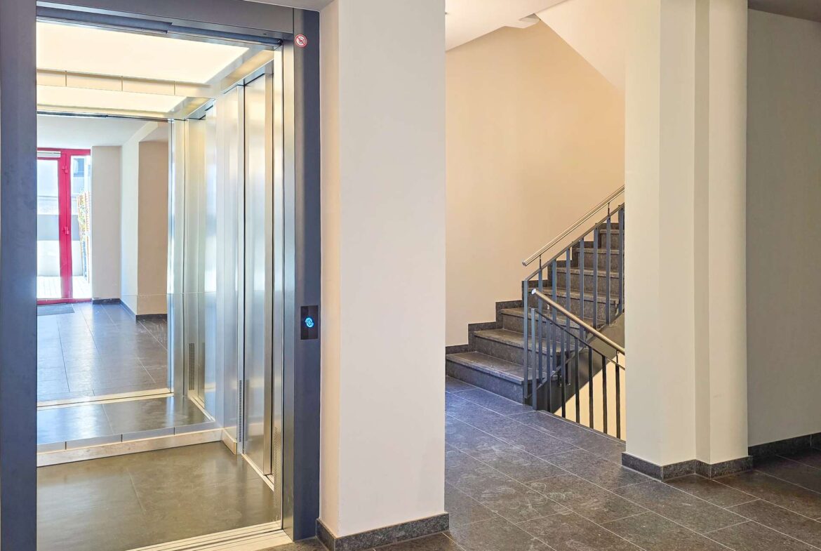 Interior lobby with an open glass-front elevator on the left and a metal staircase on the right, beige walls and dark tiled floor.