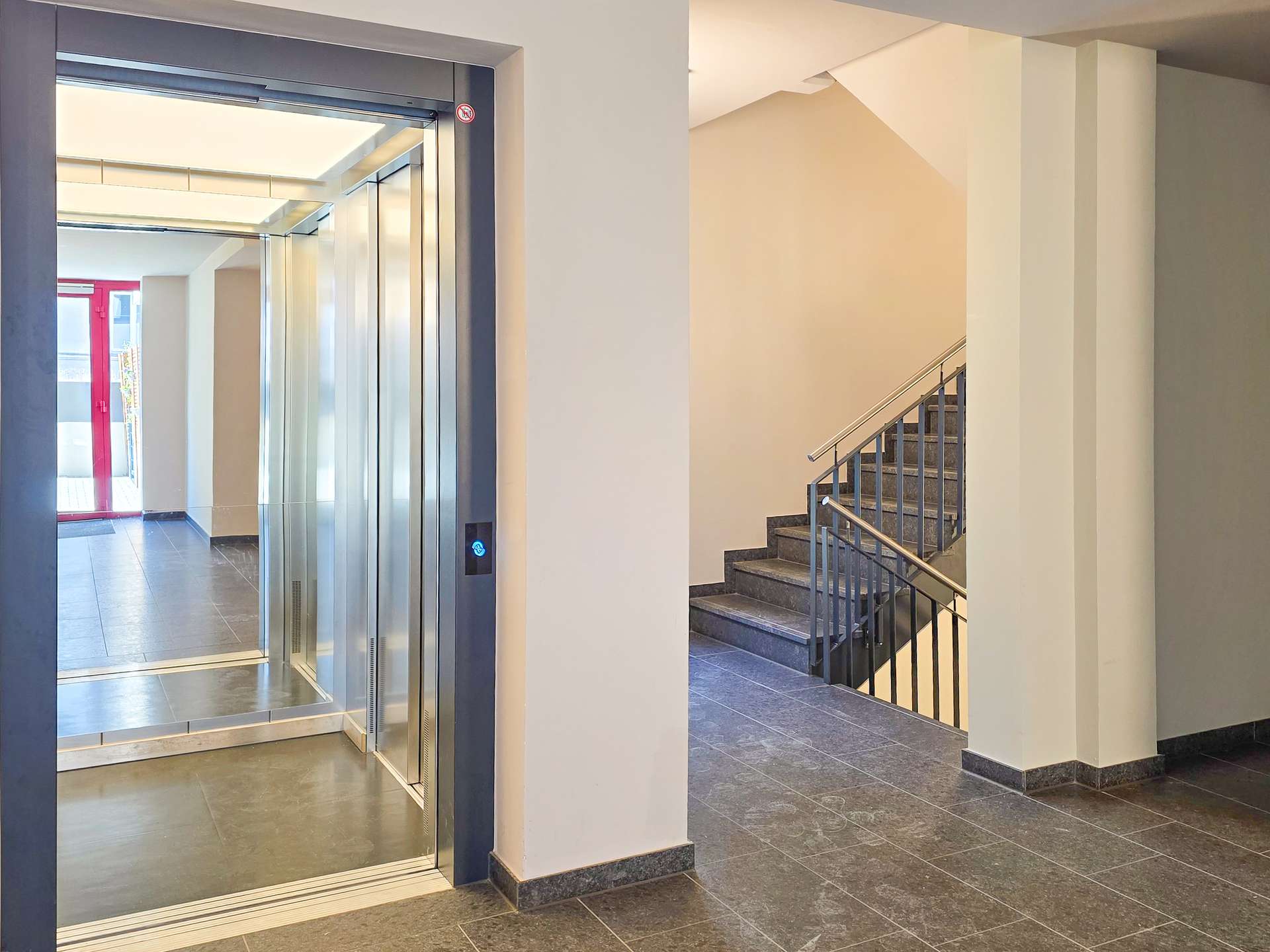 Interior lobby with an open glass-front elevator on the left and a metal staircase on the right, beige walls and dark tiled floor.