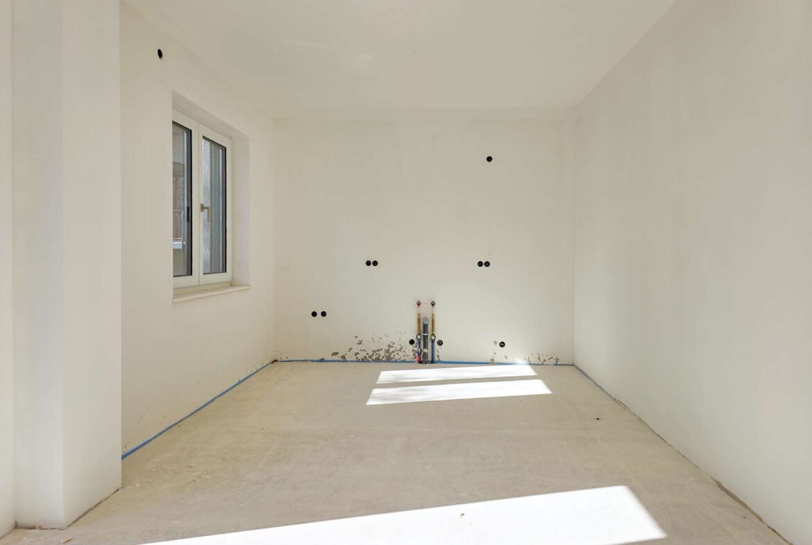 Unfinished white room with a window on the left, exposed plumbing and electrical outlets on the back wall, bare concrete floor.