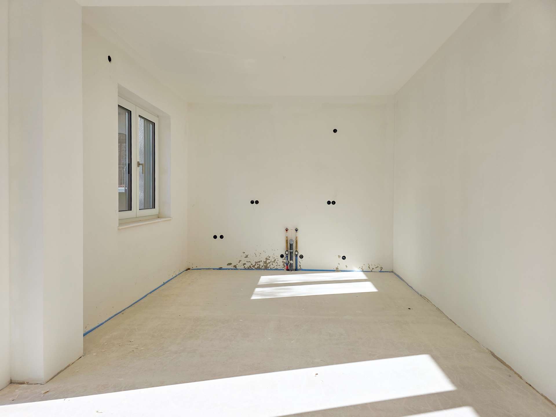 Unfinished white room with a window on the left, exposed plumbing and electrical outlets on the back wall, bare concrete floor.
