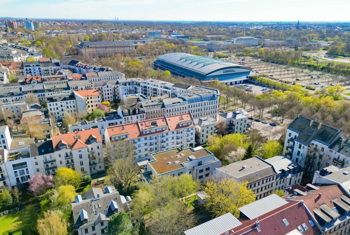 Aerial view of a city with a large curved-roof stadium amid residential buildings and spring trees under a blue sky, daytime.
