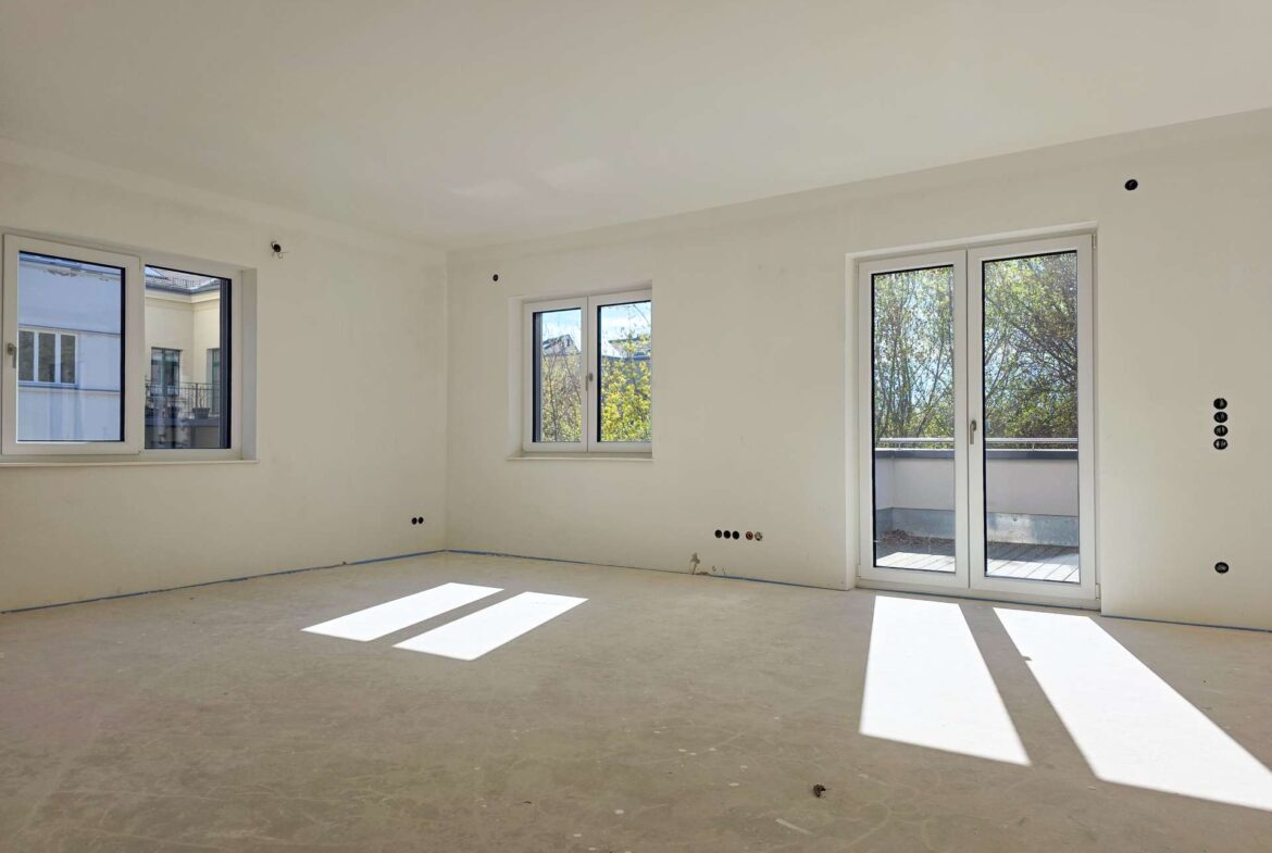 Bright, empty room under construction with white walls, two windows and a balcony door casting rectangular light patches on the floor.