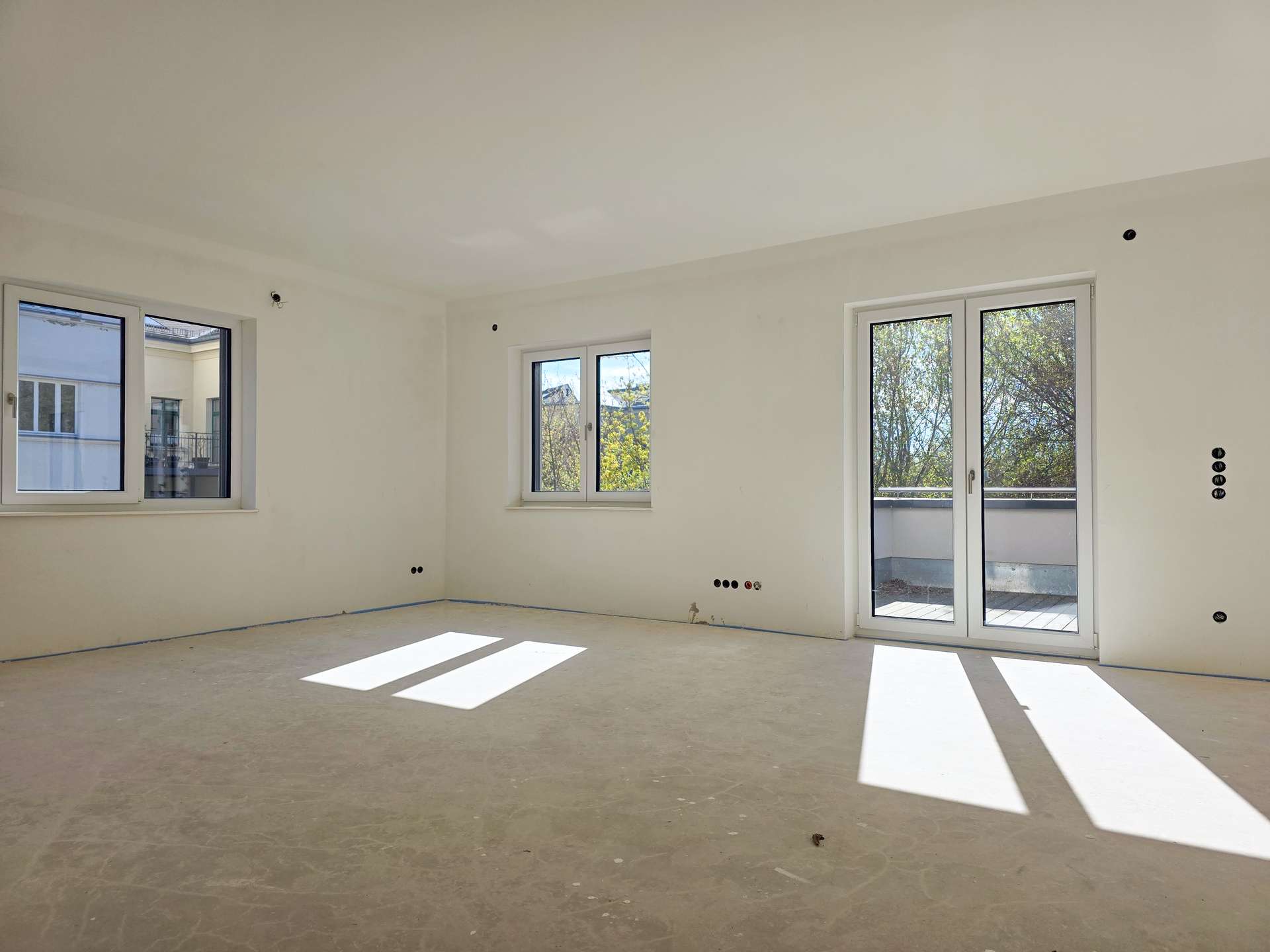 Bright, empty room under construction with white walls, two windows and a balcony door casting rectangular light patches on the floor.