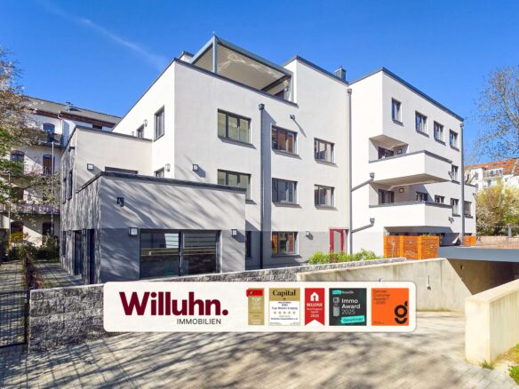 White modern apartment building with several windows and balconies under a clear blue sky; Willuhn Immobilien sign in the foreground.