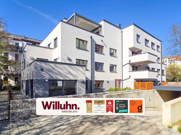 White modern apartment building with several windows and balconies under a clear blue sky; Willuhn Immobilien sign in the foreground.